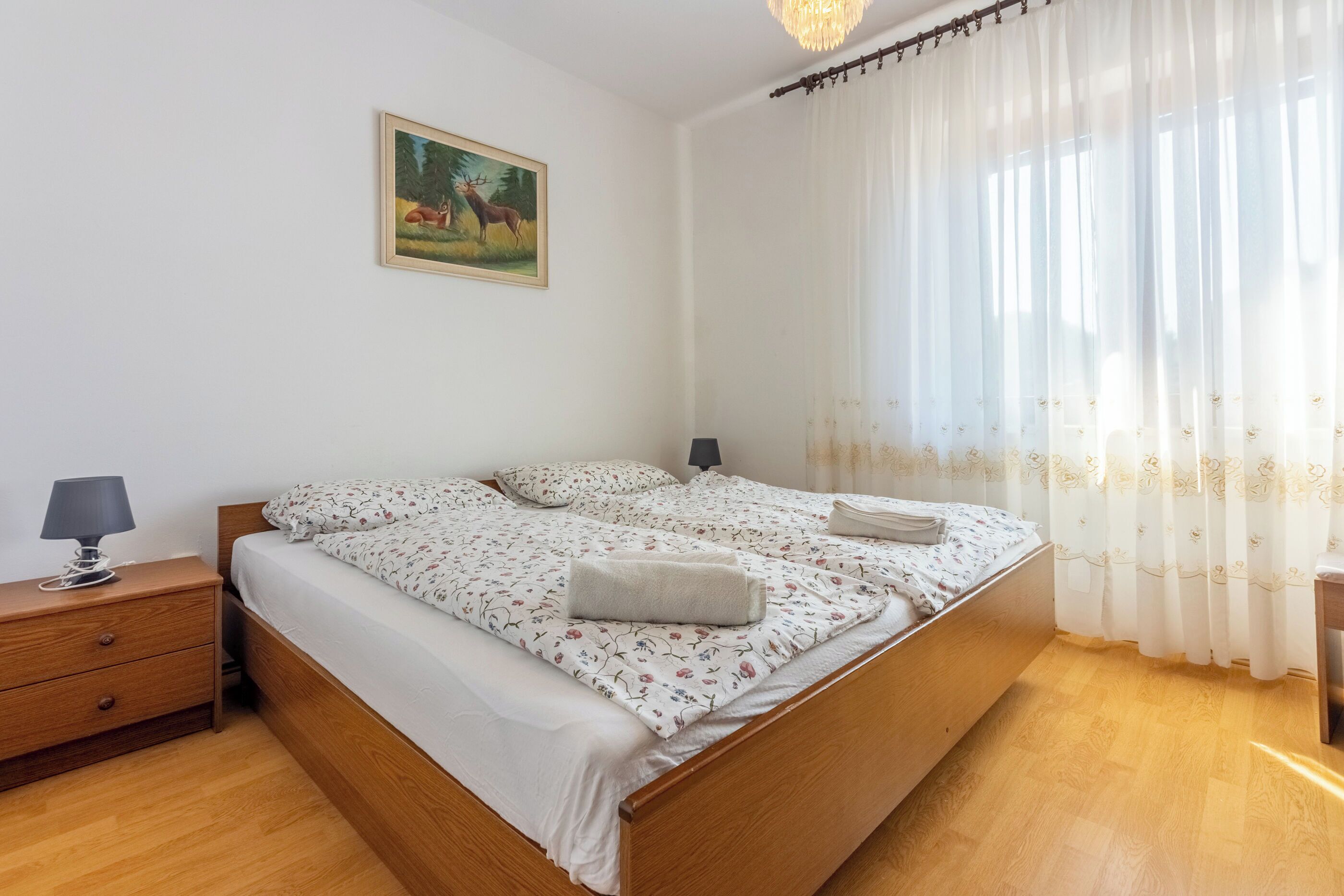Un letto rifatto con lenzuola a motivi floreali, una lampada da comodino e un quadro con cervi appeso al muro.