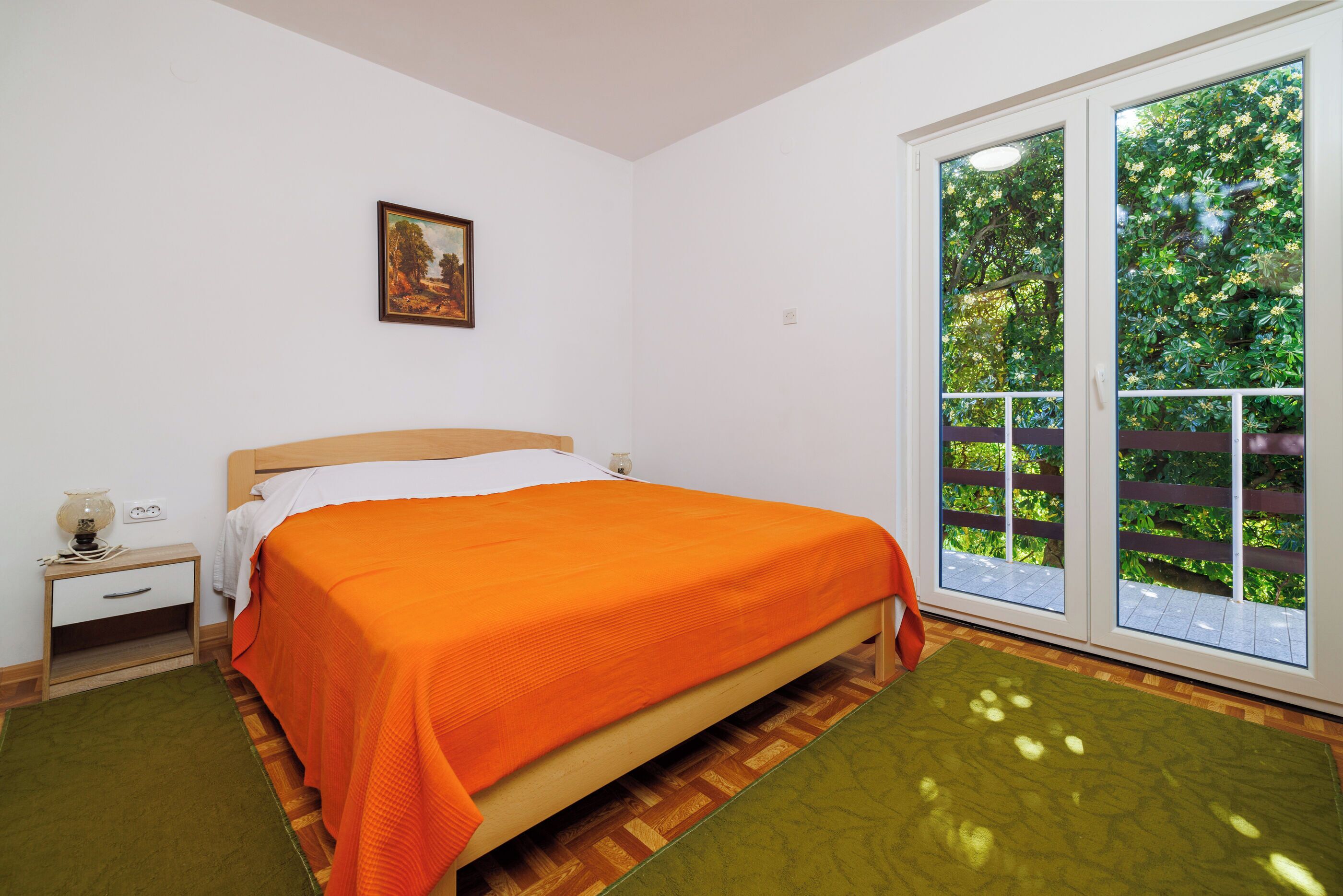 A bedroom with a bed, a nightstand, a window with a view of greenery, and a painting on the wall.