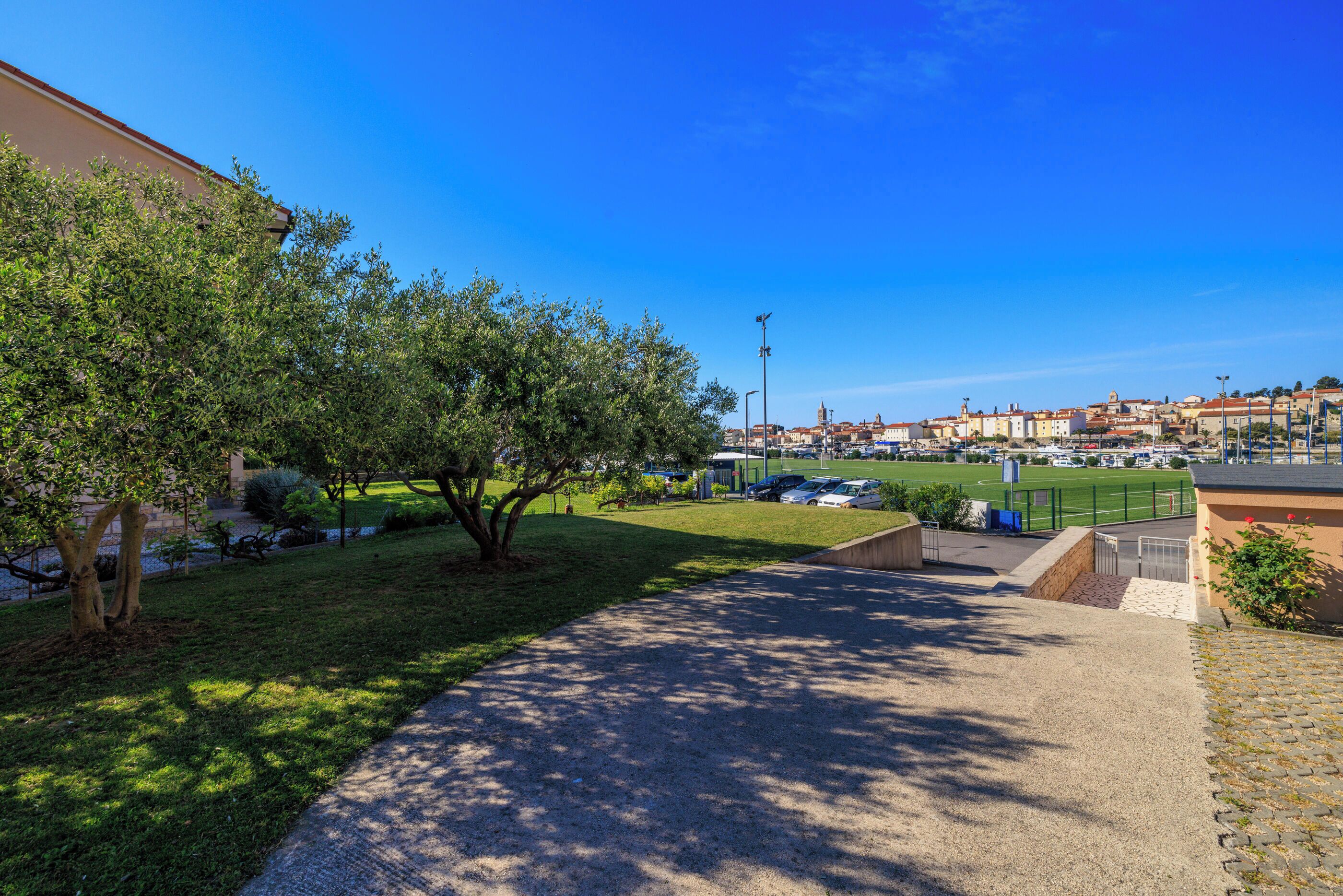 A paved pathway leading to a grassy area with trees, a sports field, and buildings in the background.