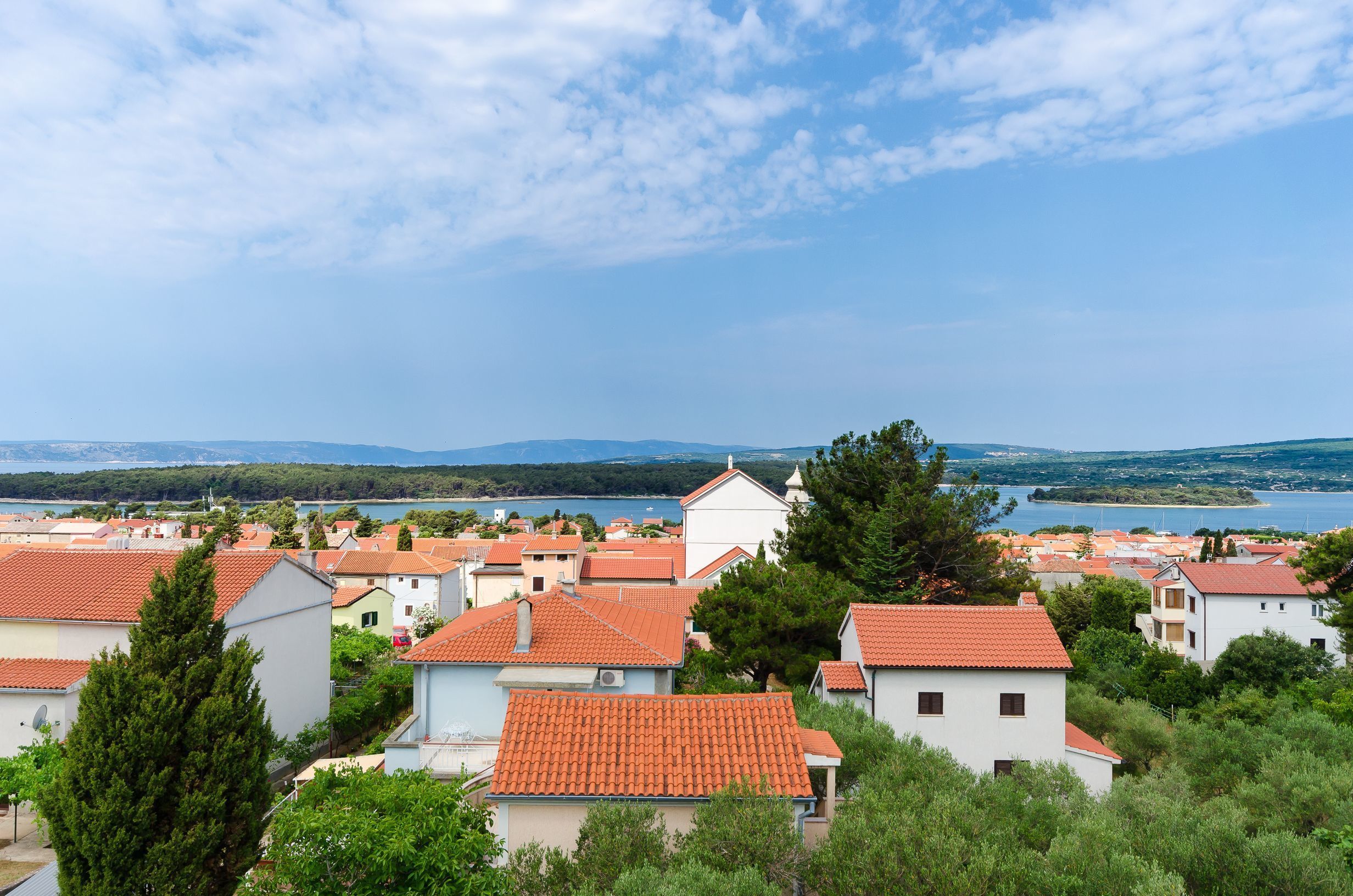 A coastal town with terracotta-roofed houses, a body of water, and distant mountains.