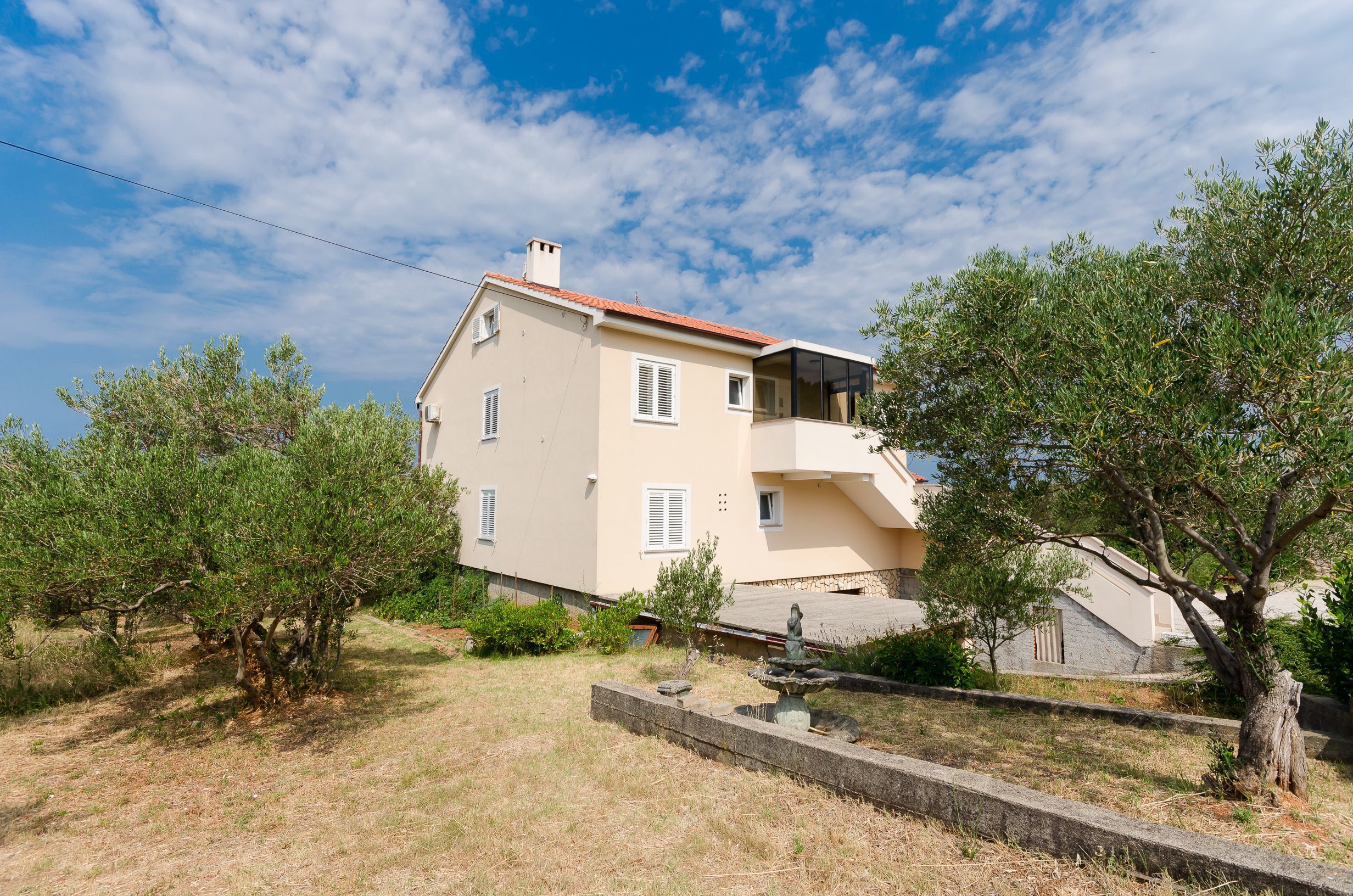 A two-story house with a red-tiled roof, a balcony, and a garden with trees and shrubs.