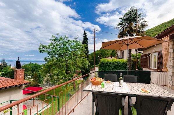 A balcony with a table set for dining, overlooking a lush garden and a distant view of the sea.