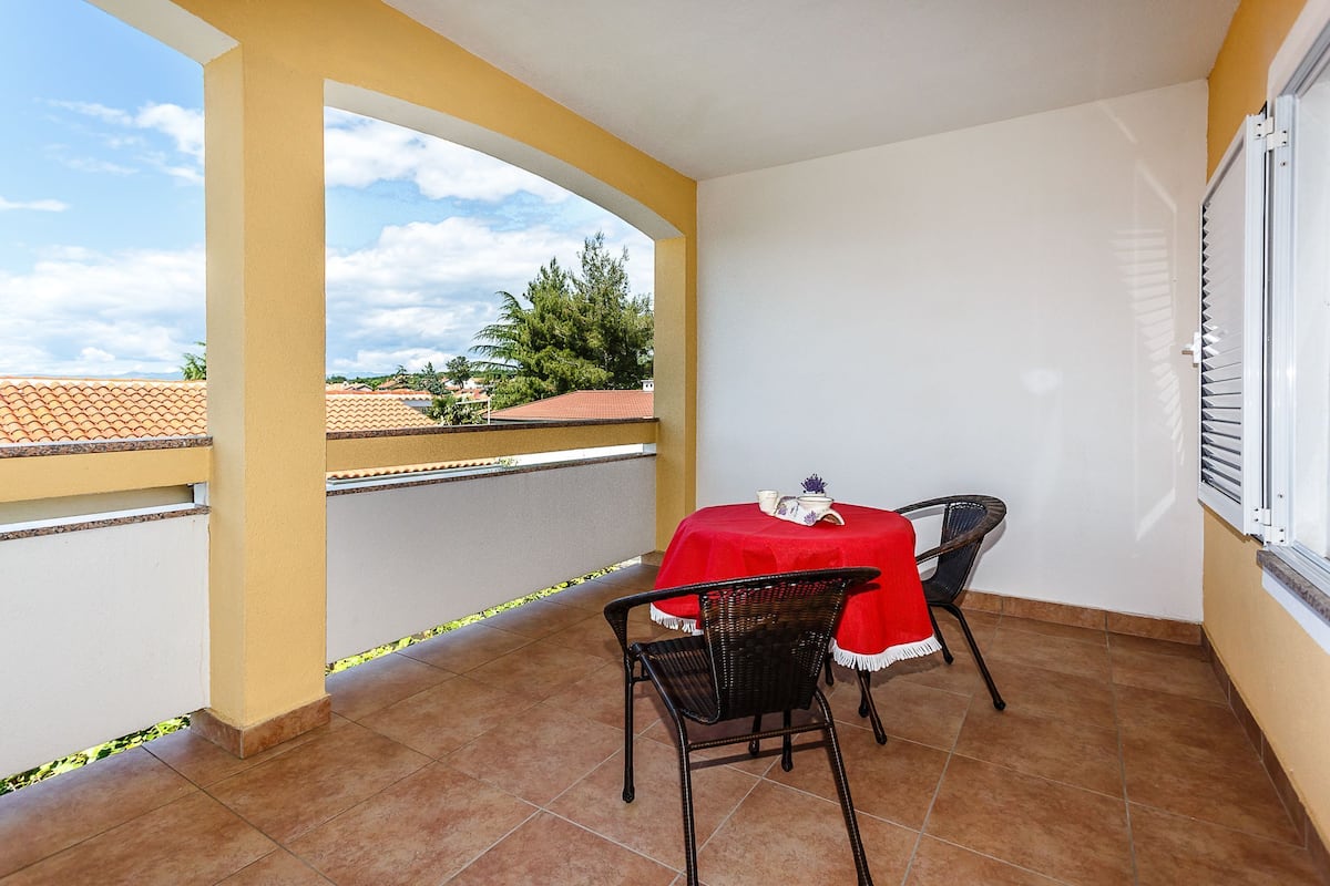 A balcony with a red table and two chairs, a yellow wall, and a view of a red-roofed building and trees.