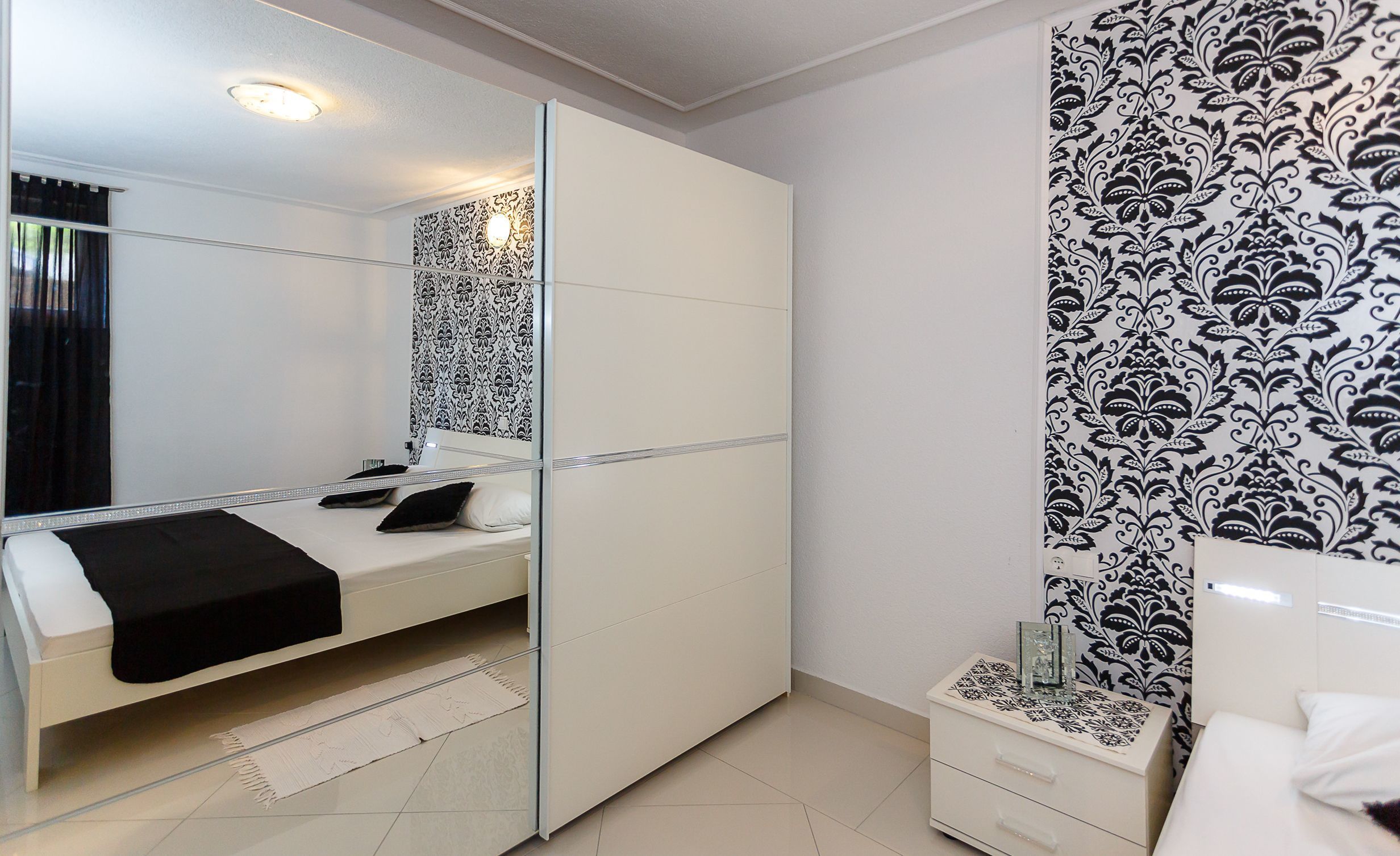 A modern bedroom with a bed, a bedside table, a mirror, and patterned wallpaper.