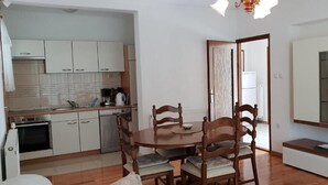 A compact kitchen with white cabinets, a wooden dining table with four chairs, and a small cabinet with a glass door. - Apartment Ljiljana (Novi Vinodolski)