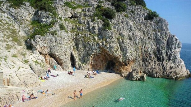 A rocky beach with clear turquoise water, a cave entrance, and people sunbathing. - Apartment Sanja (Brsec)