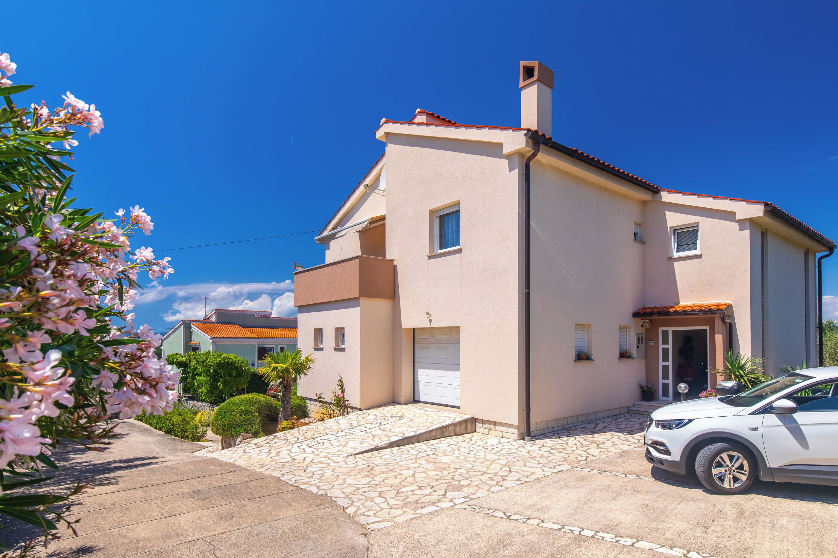 Una casa de dos pisos con camino de entrada de piedra, un coche blanco estacionado y un cielo azul despejado.
