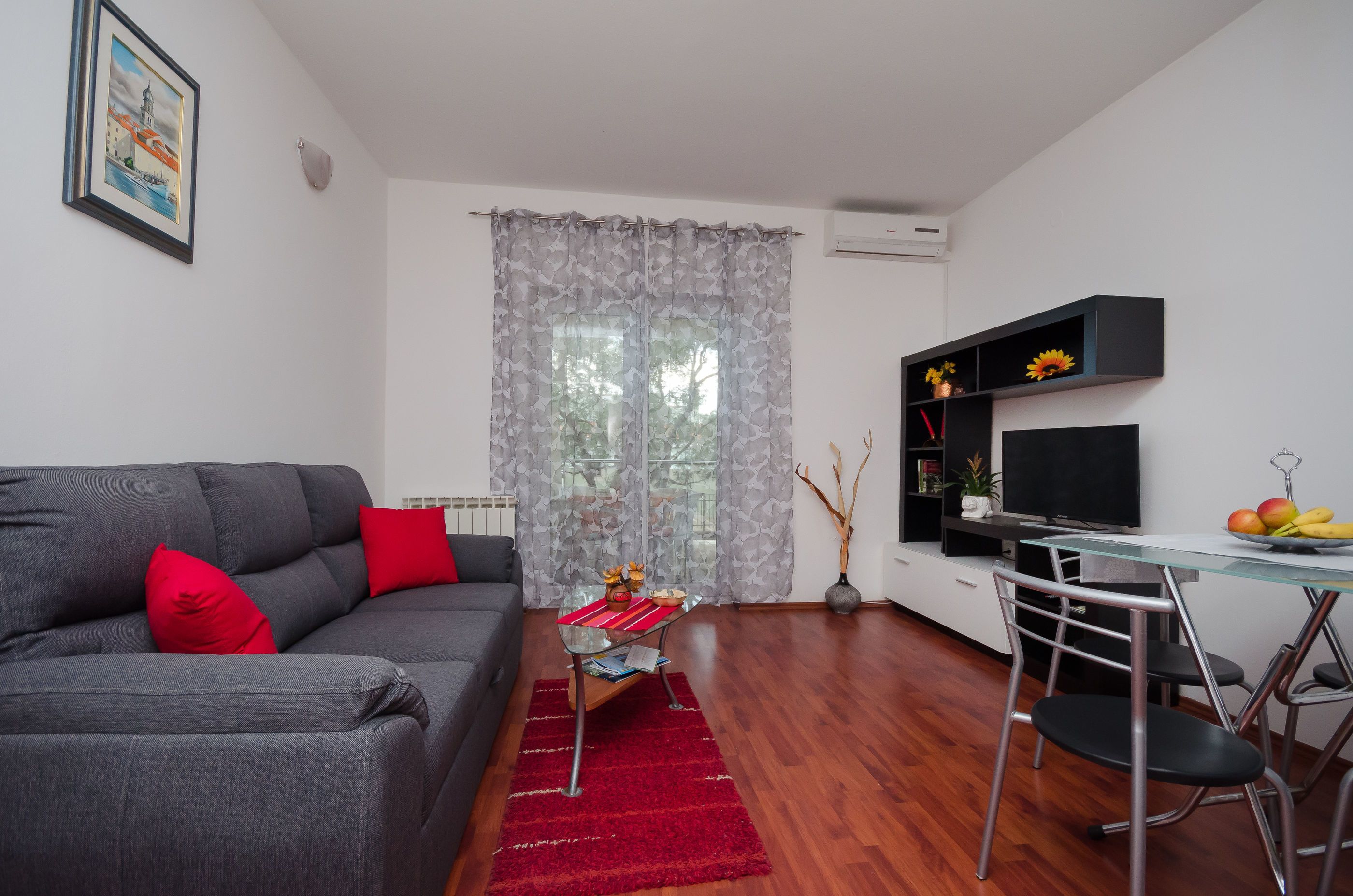 Un salon moderne avec un canapé gris, des coussins rouges, une table basse en verre et un tapis rouge. On y trouve également un coin repas avec une table et des chaises en verre, ainsi qu’un téléviseur posé sur un meuble noir.