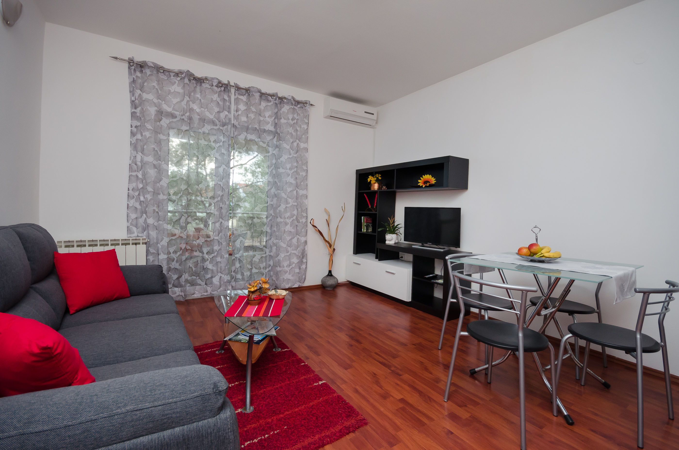 A modern living room with a grey sofa, red cushions, a glass coffee table, and a dining area with a glass table and metal chairs.