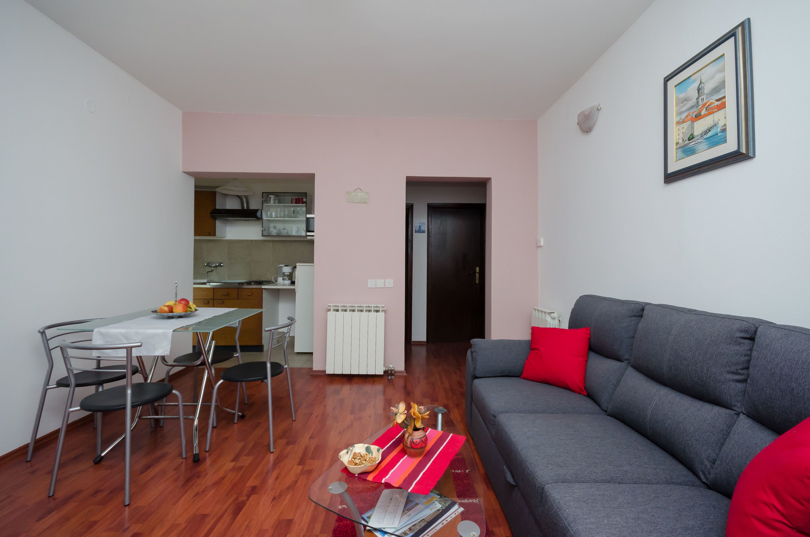 Un salon avec un canapé gris, une table à manger avec des chaises, une cuisine en arrière-plan et un tableau encadré au mur.
