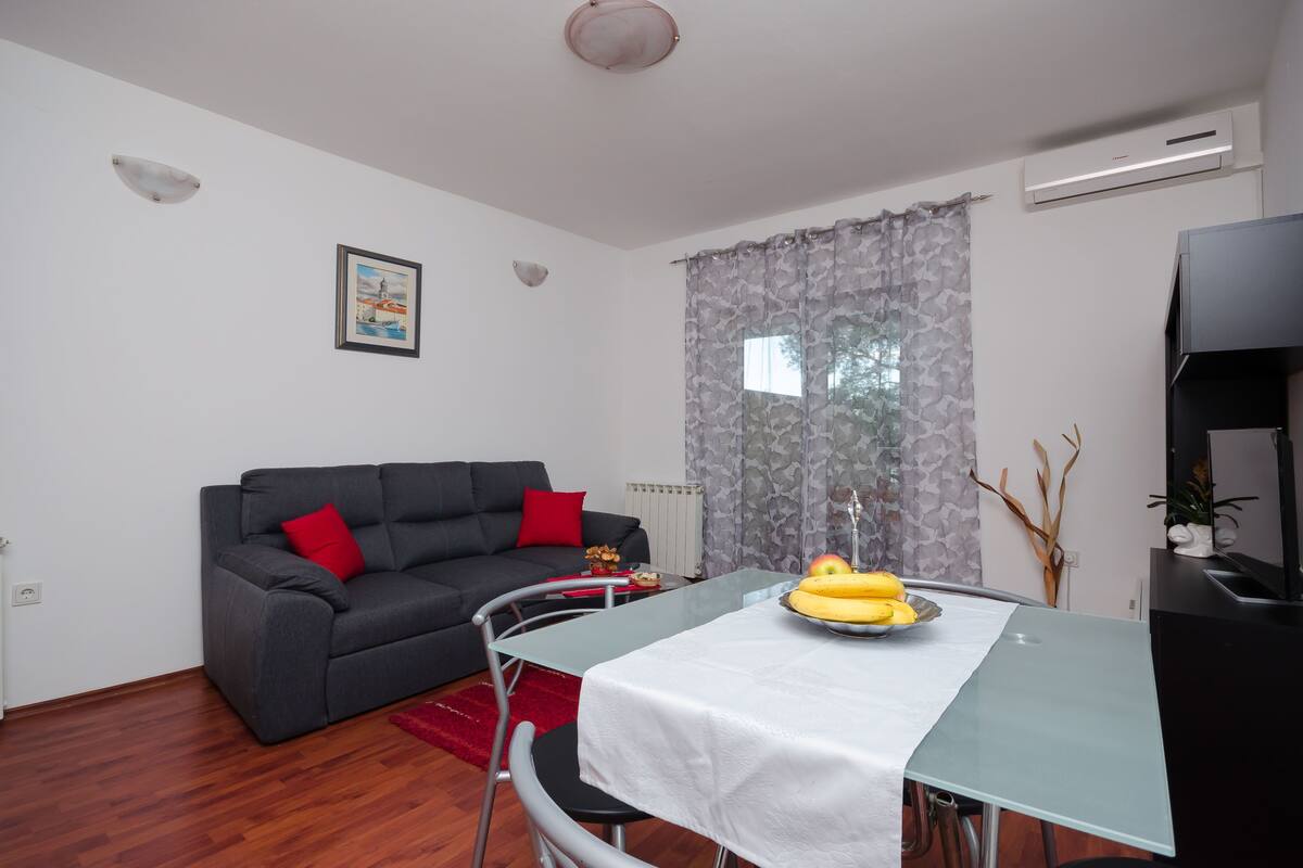 Un salon avec un canapé gris, une table à manger avec un plat décoré de motifs représentant des bananes, un radiateur et un rideau gris.