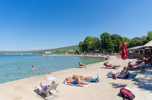 A beachside setting with sun loungers, umbrellas, and people relaxing by the water. - Apartments Miljenko (Punat)