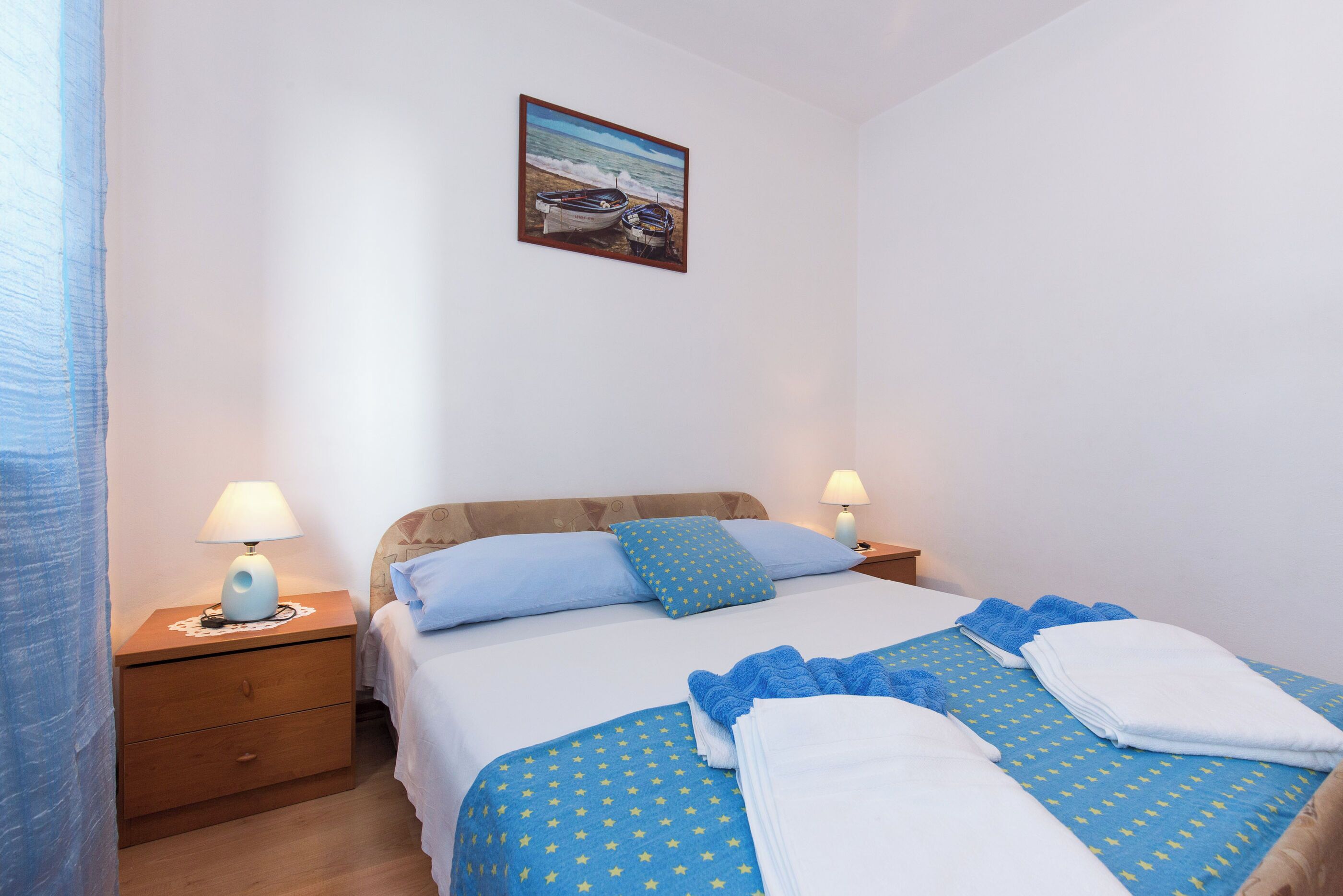 A neatly made bed with blue and white bedding, two bedside tables with lamps, and a painting of a boat on the wall.