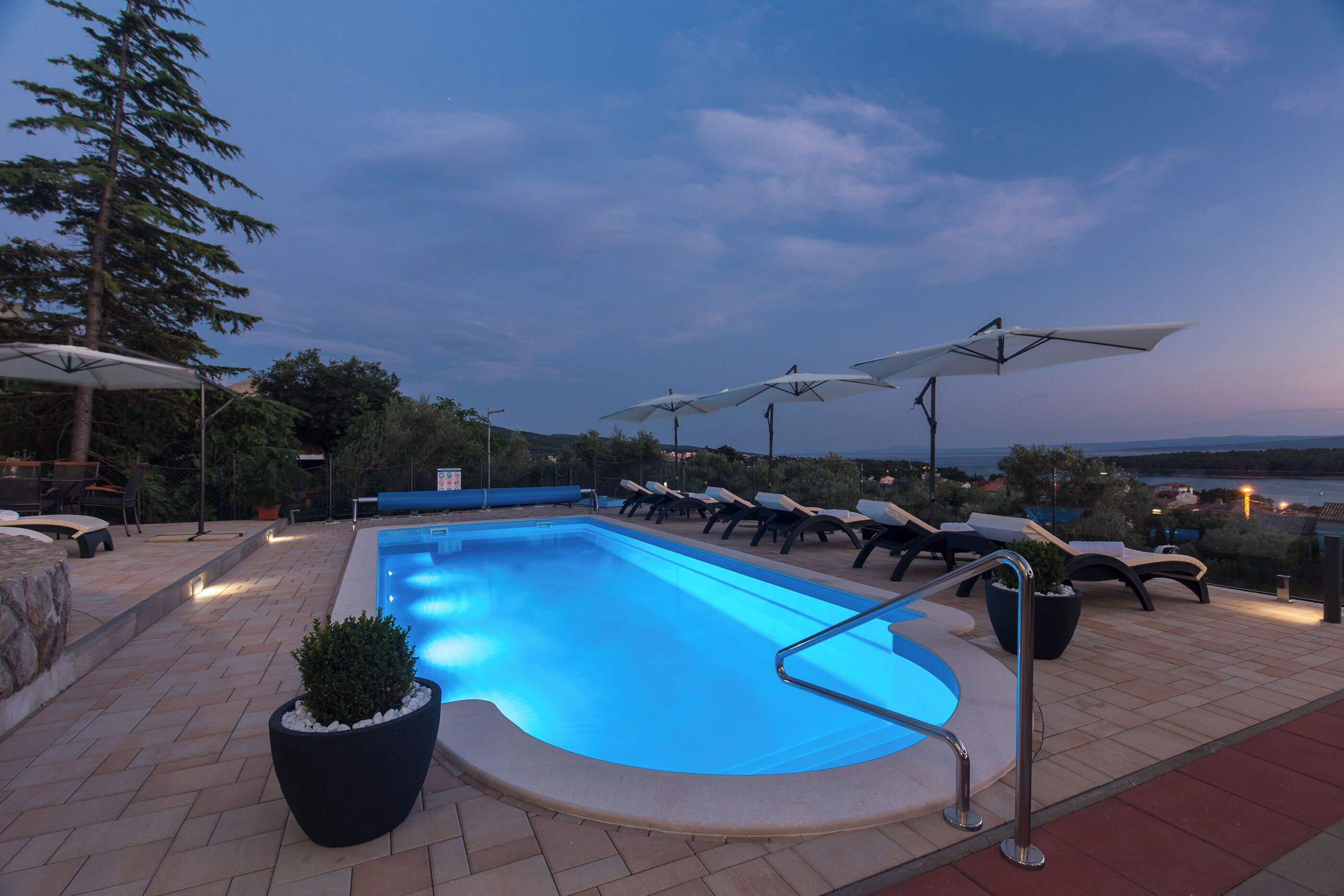 A swimming pool with lounge chairs and umbrellas, surrounded by potted plants and a view of the landscape.