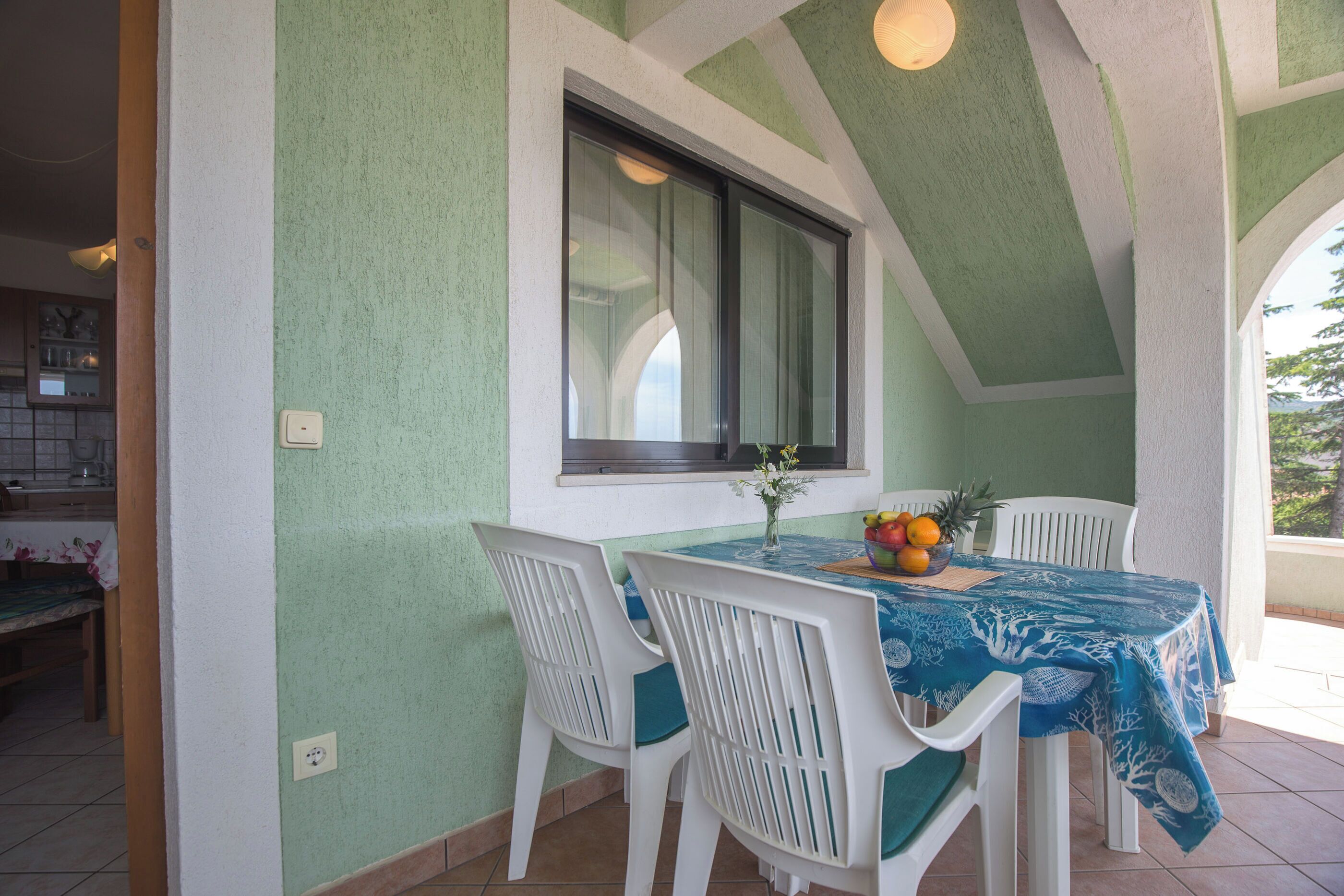 A dining area with a table covered by a blue tablecloth, white chairs, and a window with curtains.