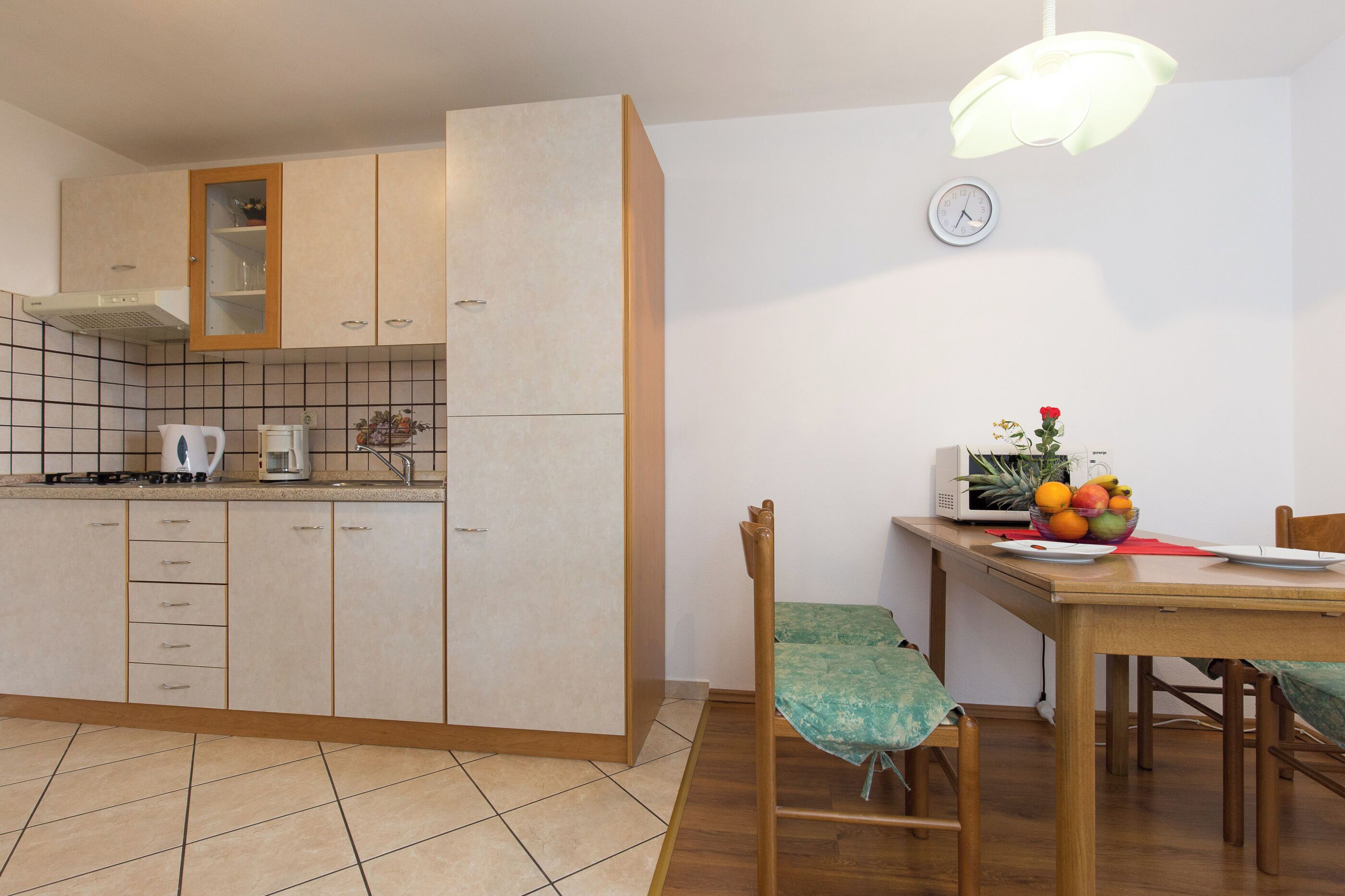 A kitchen with a microwave, kettle, and a fruit bowl on the counter. There is a dining table with chairs and a clock on the wall.