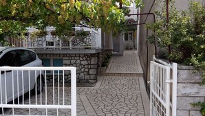 A stone pathway leading to a gated entrance, with a parked car and a white gate in the foreground. - Apartments Jasminka (Selce)