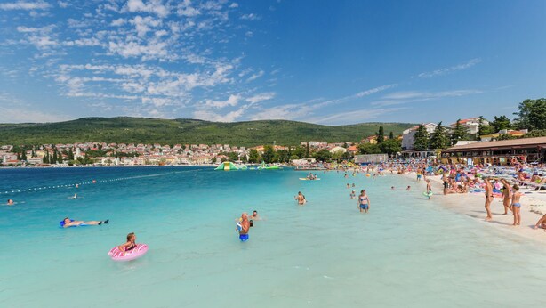 A beach with clear turquoise water, people swimming and lounging, and a distant view of a town with buildings and hills. - Apartments Jasminka (Selce)