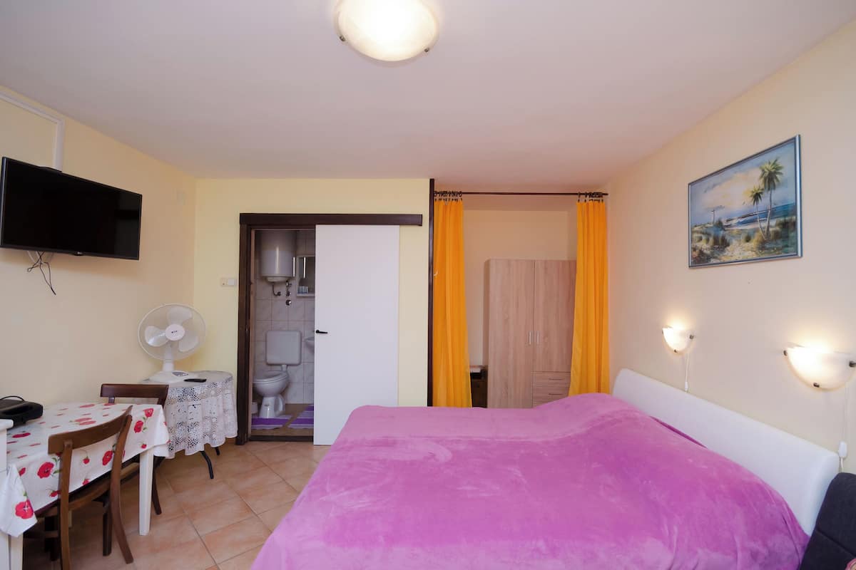 A bedroom with a bed, a TV, a desk with a chair, and a bathroom visible through an open door.