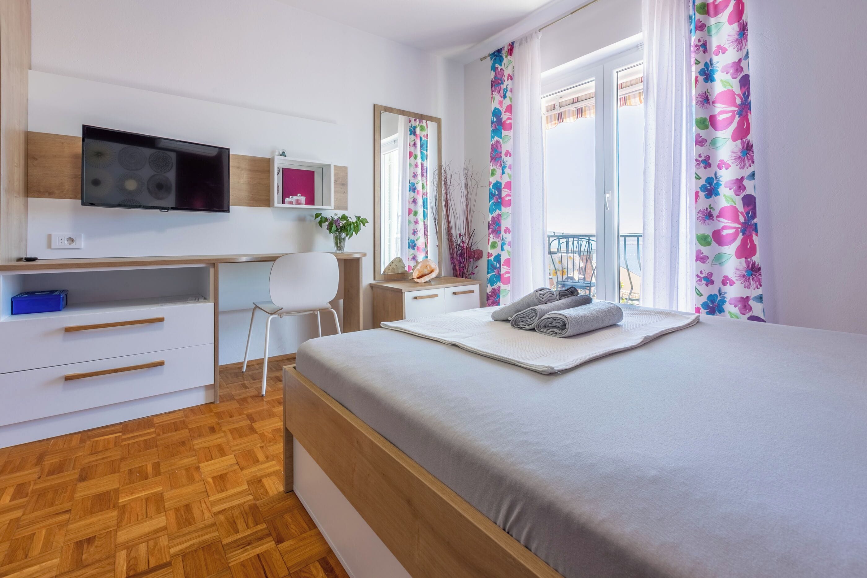 A bedroom with a bed, a desk, a chair, a TV, and a window with curtains.