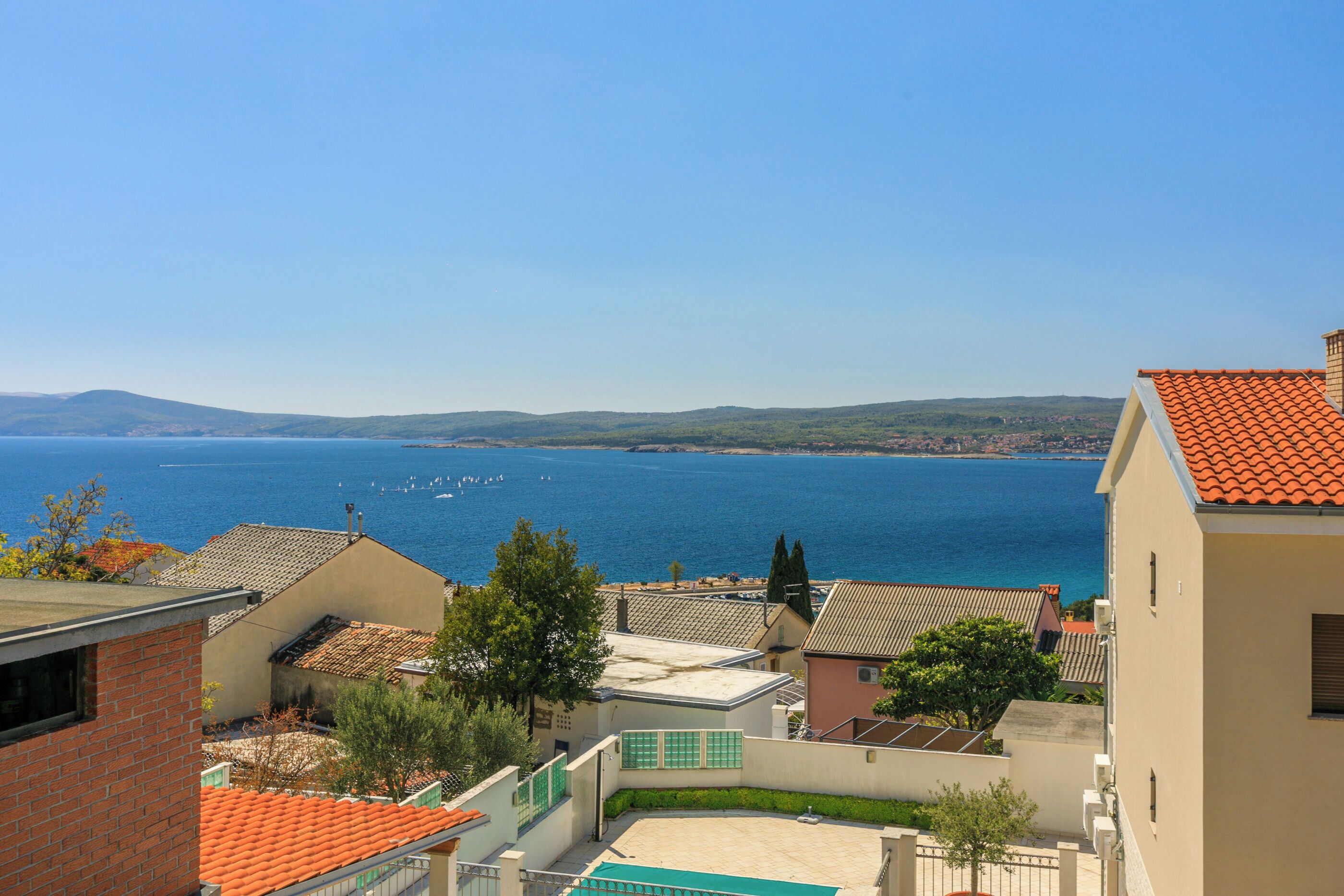 A coastal residential area with houses, a clear blue sea, and distant hills.