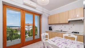 A kitchen with wooden cabinets, a dining table with a purple tablecloth, and a balcony with a view of the city. - Apartments Dijana (Crikvenica)