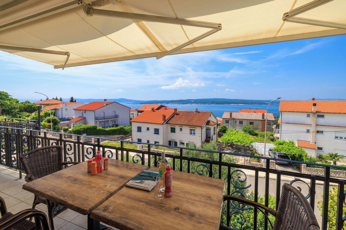 A terrace with a wooden table, wicker chairs, and a view of buildings and water.
