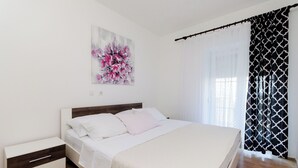 A bedroom with a bed, a nightstand, a wall-mounted painting, and a window with curtains. - Apartment Marica (Lopar)