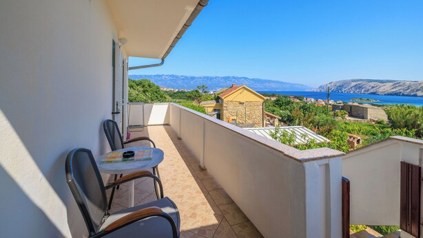 Ein Balkon mit Tisch und Stühlen, der auf eine Küstenlandschaft mit Bergen und blauem Himmel blickt.