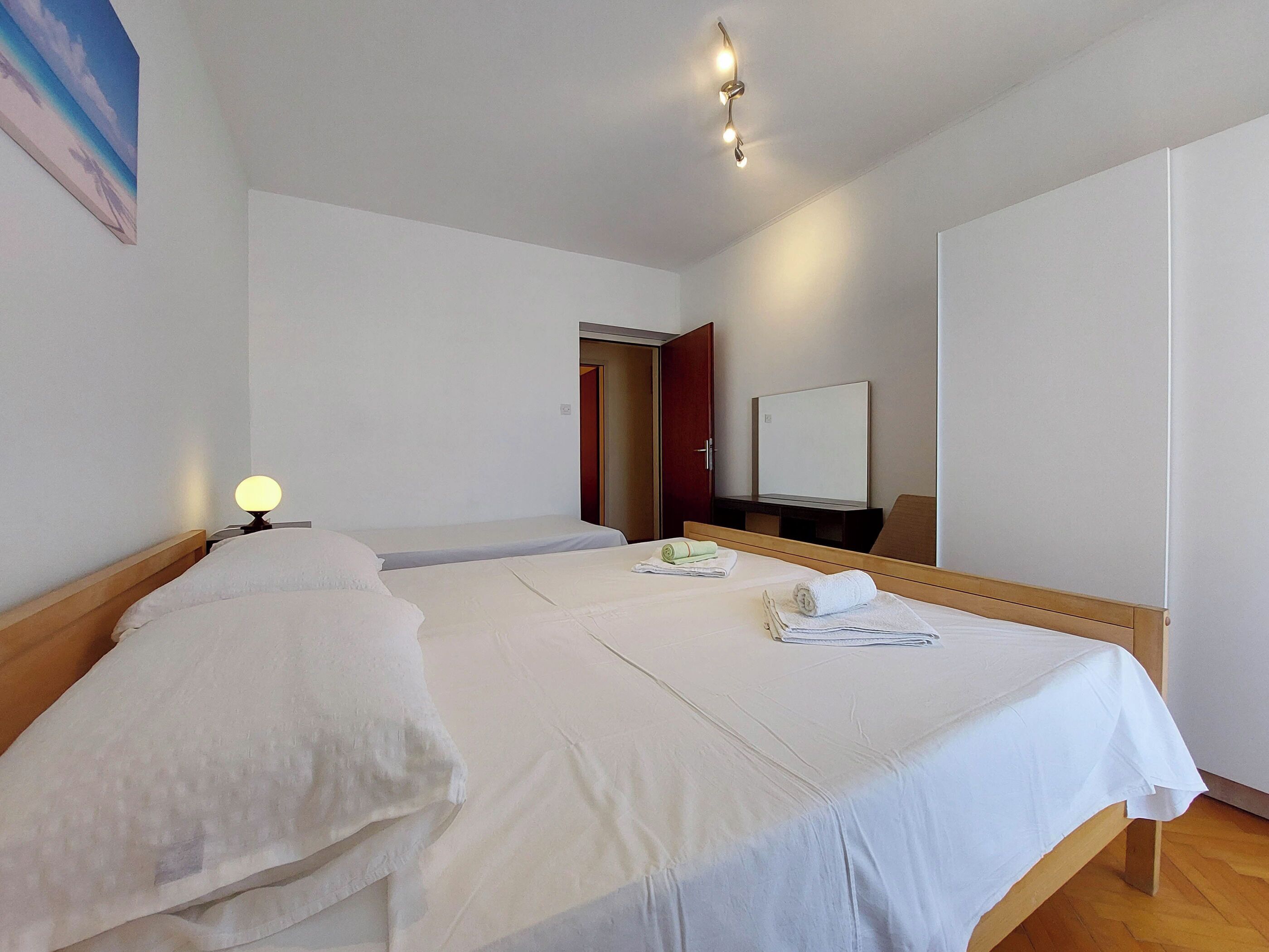 A hotel room with two single beds, a bedside table with a lamp, a wooden headboard, a mirror, and a door leading to another room.