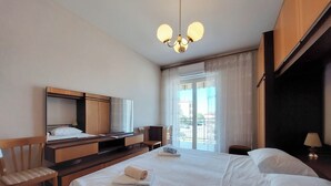 A hotel room with a bed, a desk with a chair, a TV, and a balcony with curtains. - Apartments Nikola (Punat)