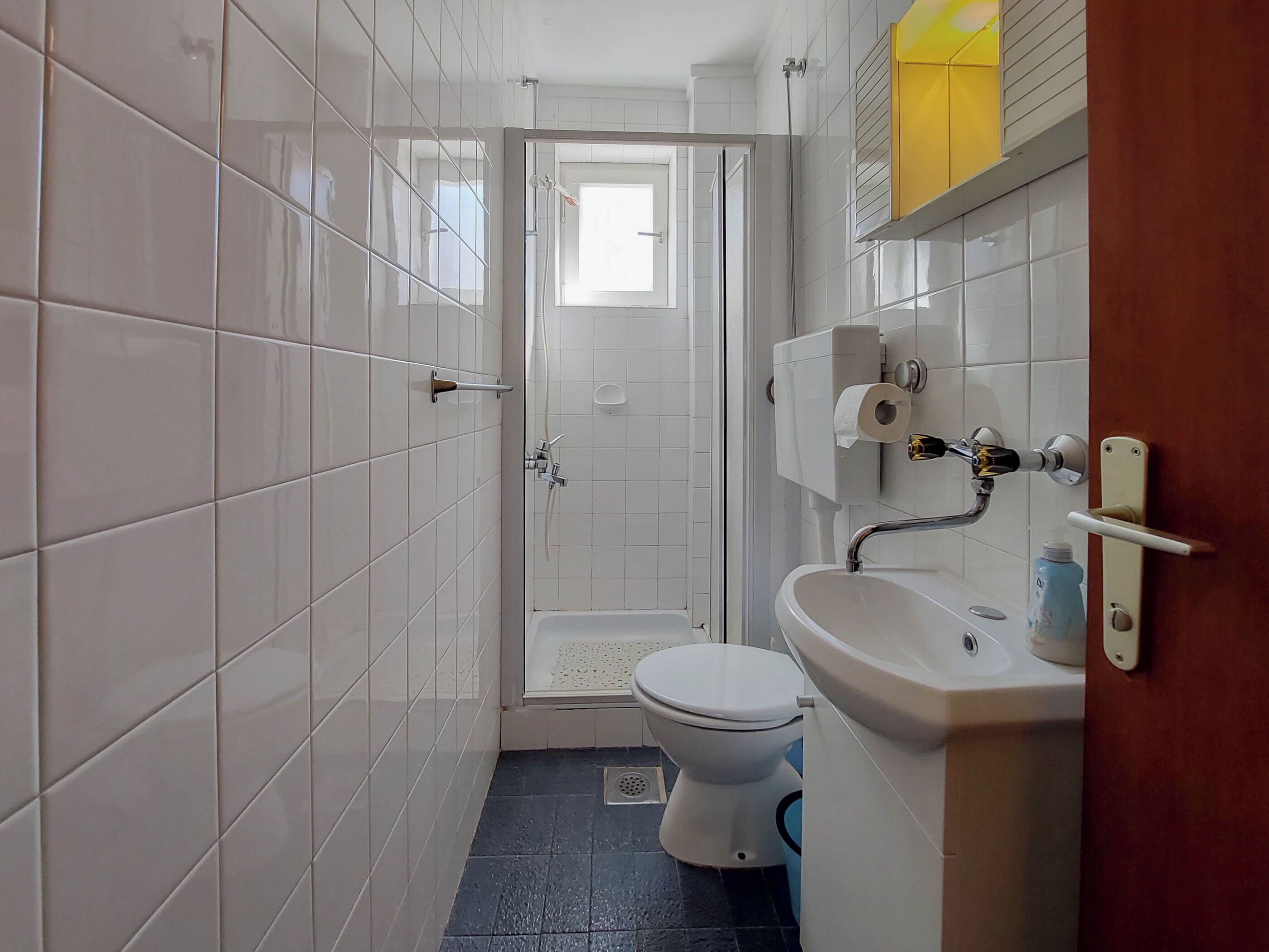 A small bathroom with white tiles, a shower, a toilet, and a sink.