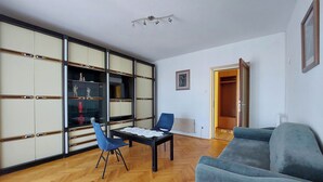 A living room with a blue sofa, a blue chair, a wooden herringbone floor, a black table with a lace tablecloth, and a sliding glass door leading to a closet. - Apartments Nikola (Punat)