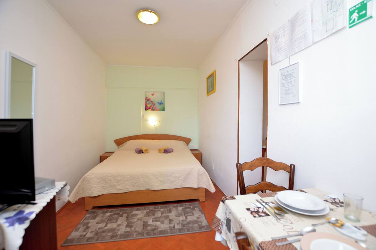 A small, well-lit bedroom with a bed, a desk with a computer, and a dining area with plates and cutlery.