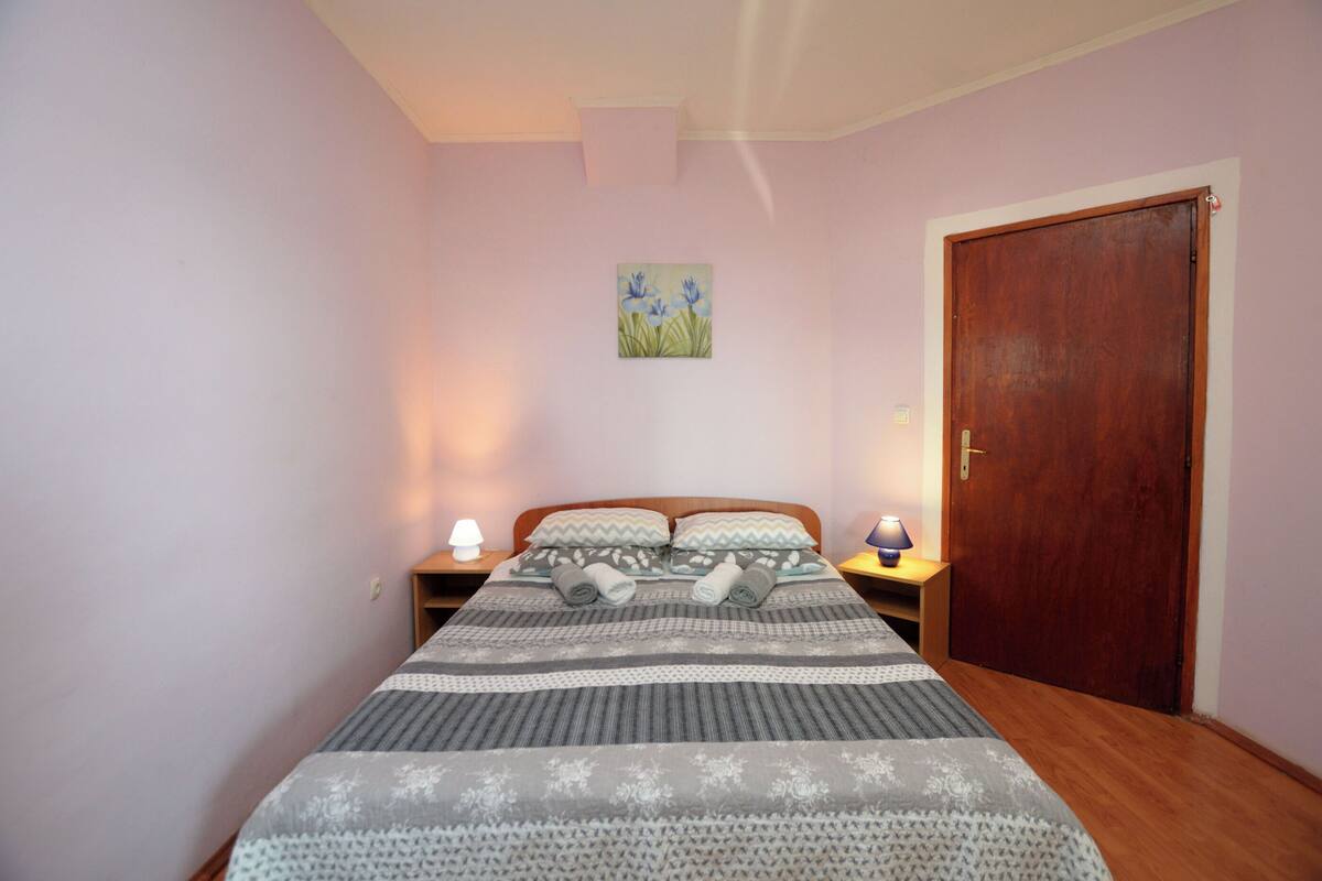 A bedroom with a bed, two bedside lamps, a wooden door, and a wall painting.