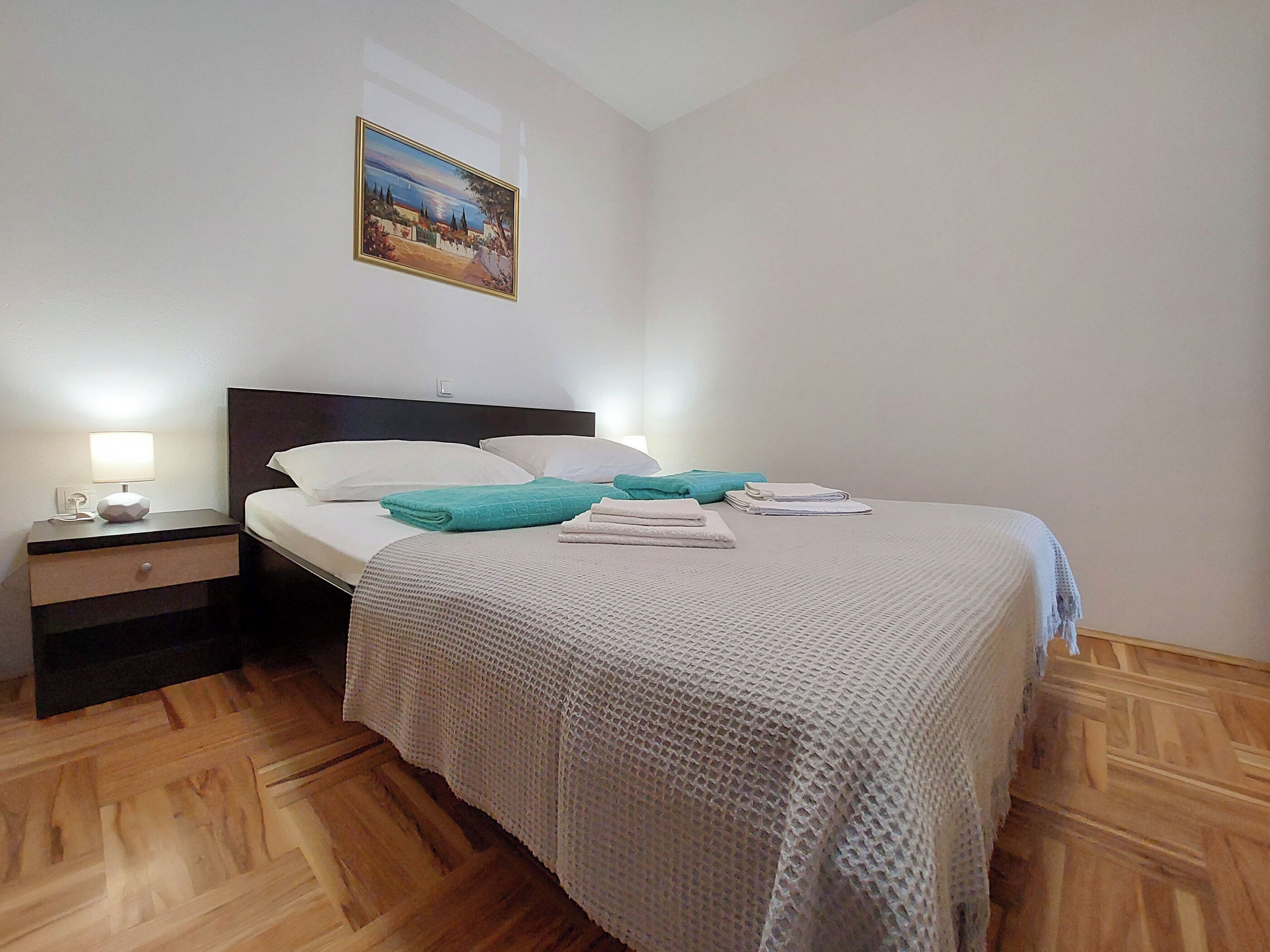 Una cama bien hecha con una manta blanca, una toalla color turquesa y una almohada blanca. Se ve una mesita de noche con una lámpara y un cajón pequeño.