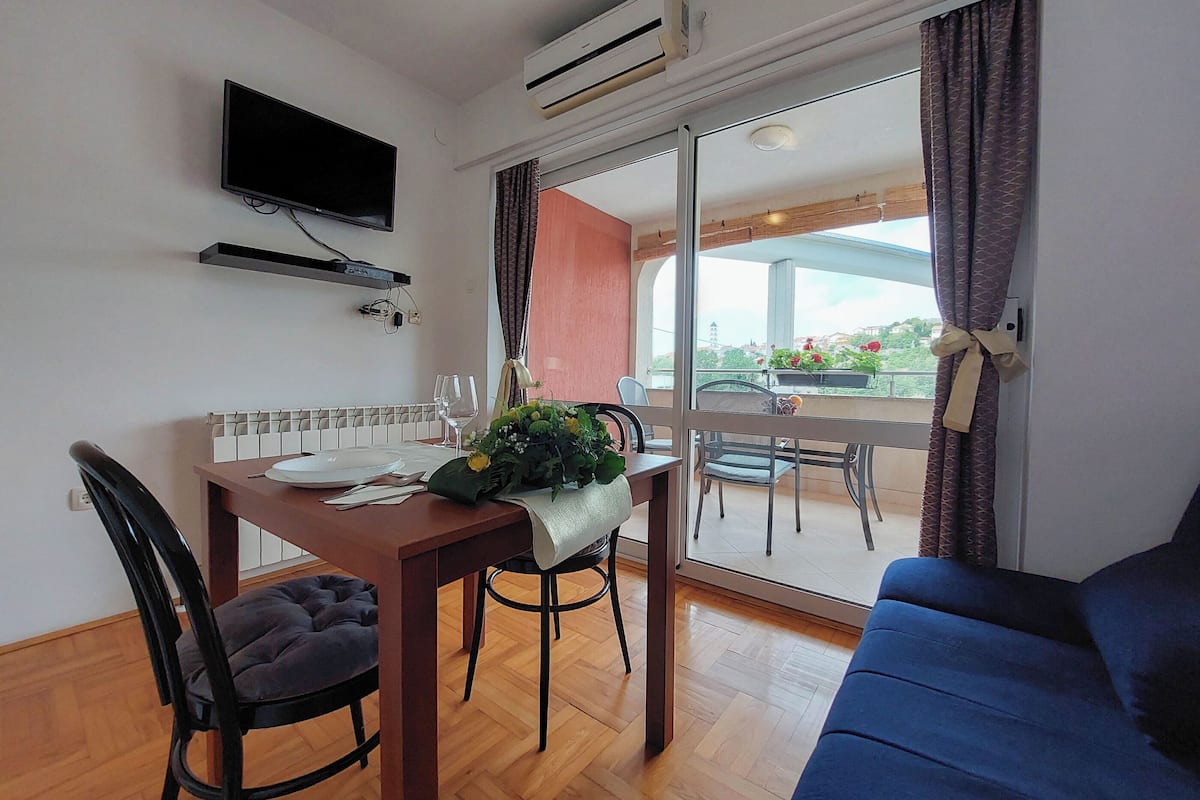 A dining area with a table set for two, a blue sofa, and a TV mounted on the wall.