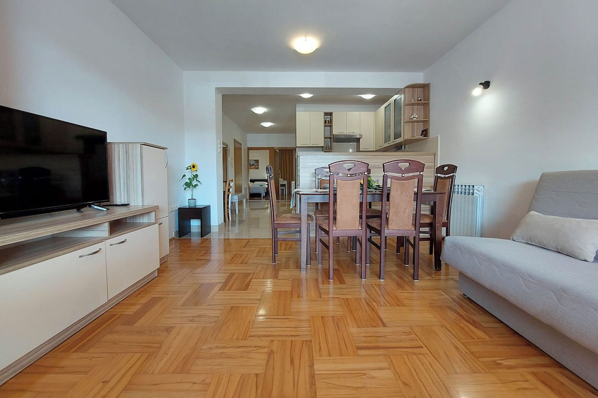 A modern living room with a flat-screen TV, wooden flooring, a sofa, and a dining area with chairs and a table.