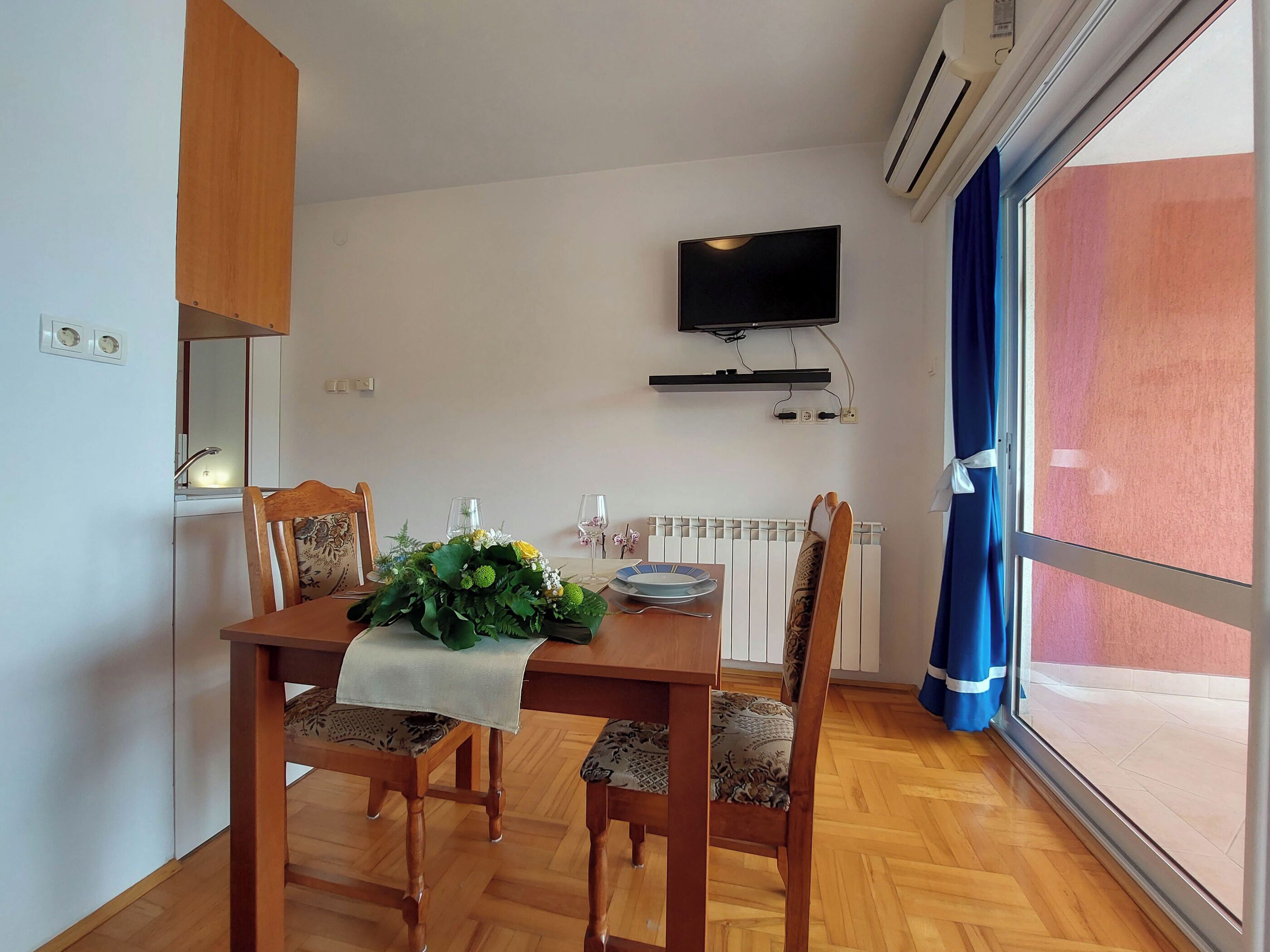 A dining area with a wooden table, four chairs, a TV mounted on the wall, and a window with curtains.