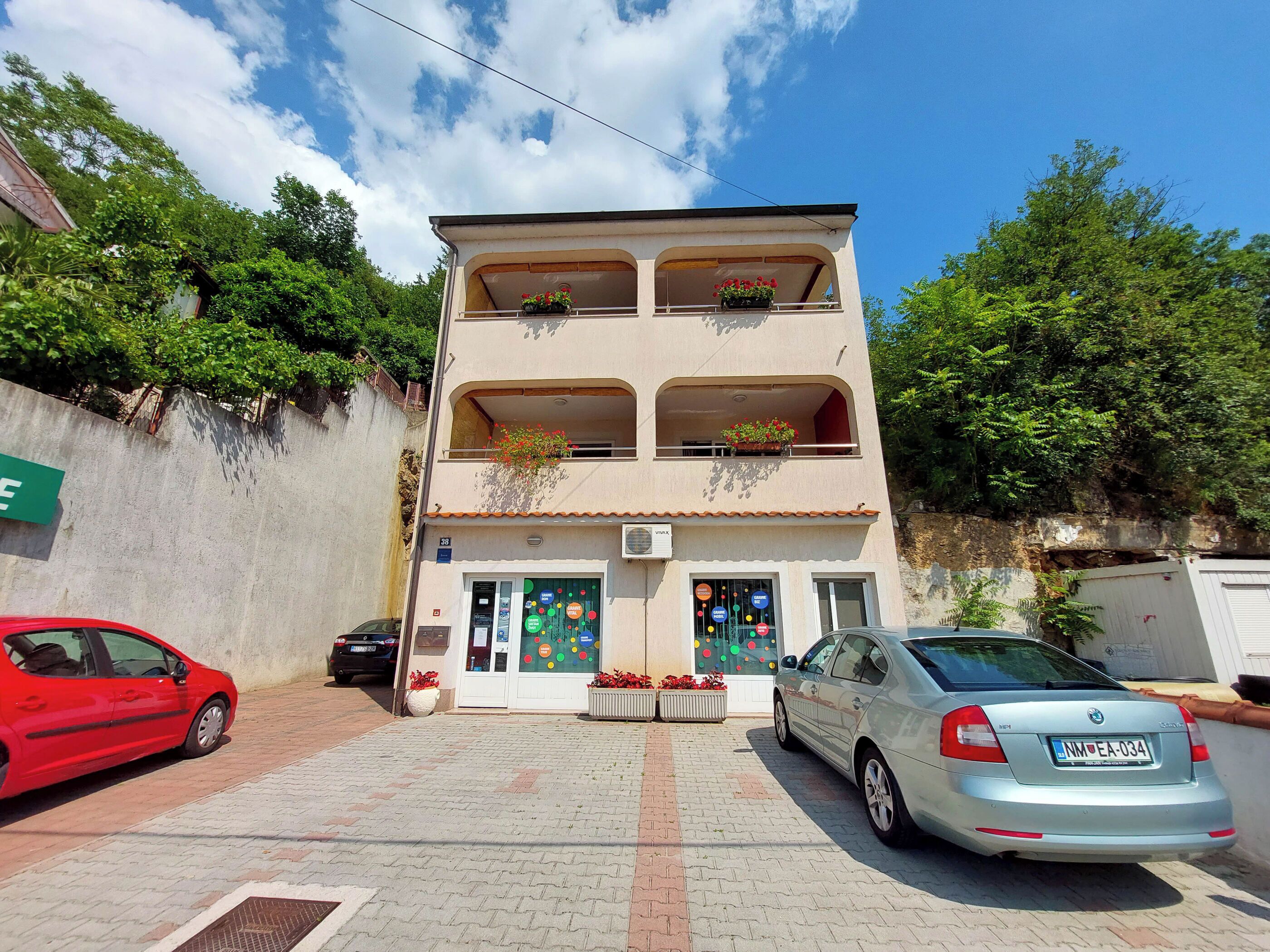 Un edificio de dos pisos con un balcón en el segundo nivel, acceso a nivel de calle y un estacionamiento con dos coches.