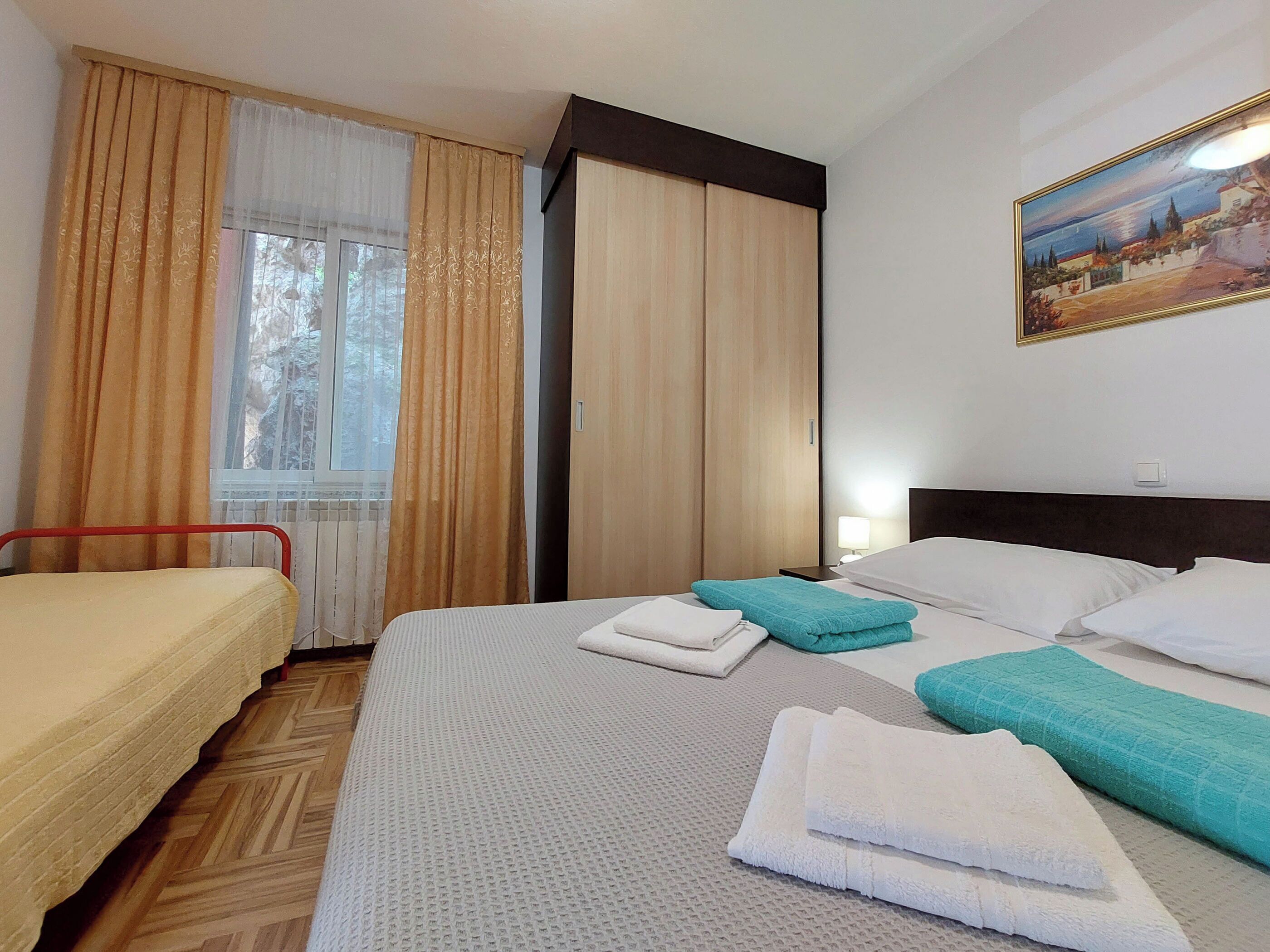 A hotel room with a bed, a chair, a window with curtains, a painting on the wall, and a radiator.