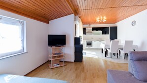 Uma sala de estar moderna com televisão, teto de madeira e uma área de jantar com cadeiras brancas e uma mesa.