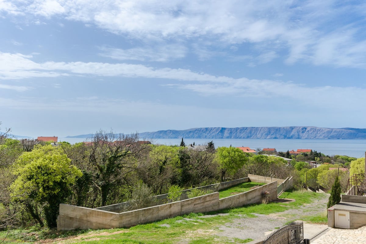 A coastal landscape with a view of the sea and mountains. There are trees and a concrete structure in the foreground.