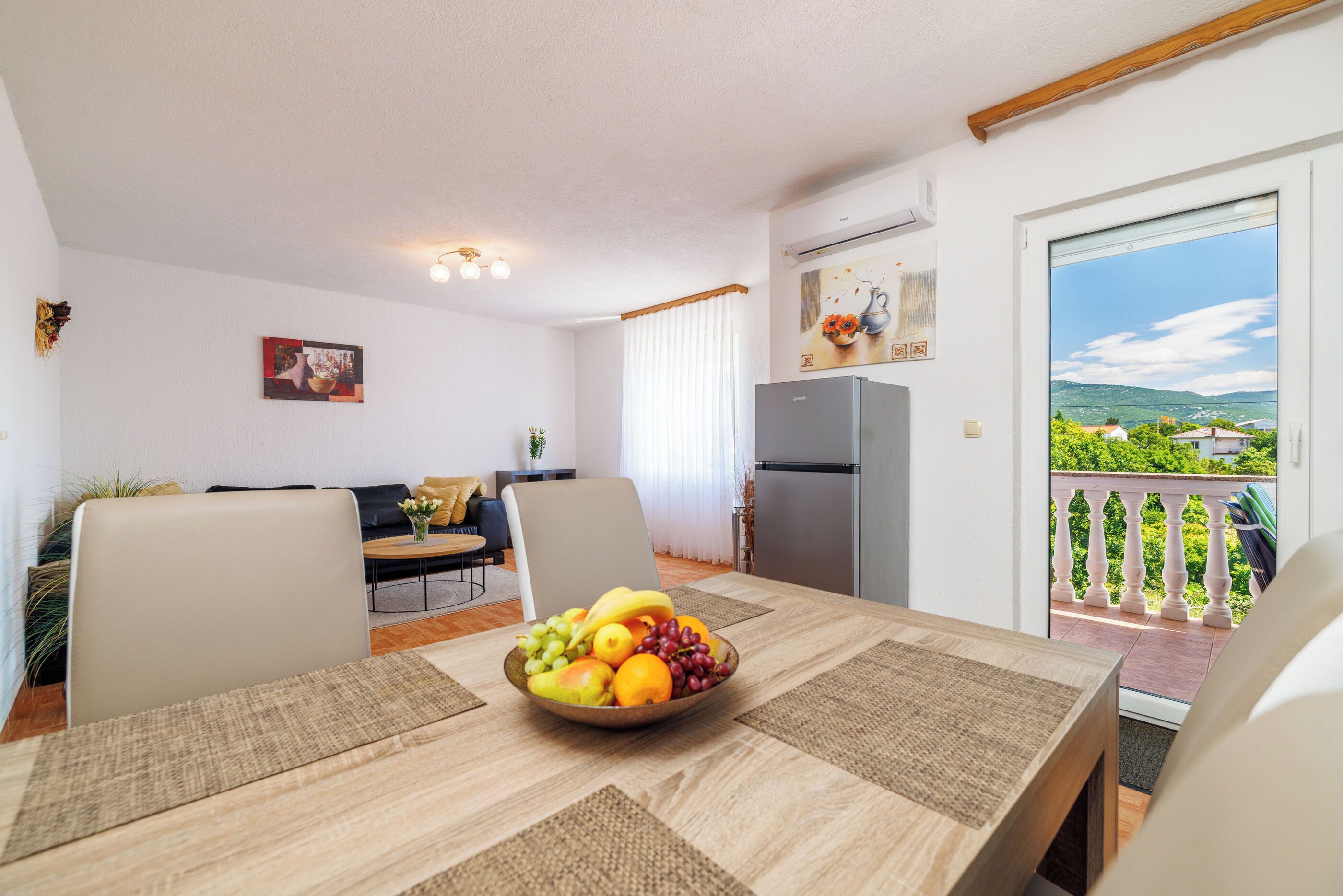 A dining area with a table set for two, a fruit bowl, and a view of a balcony with a railing.
