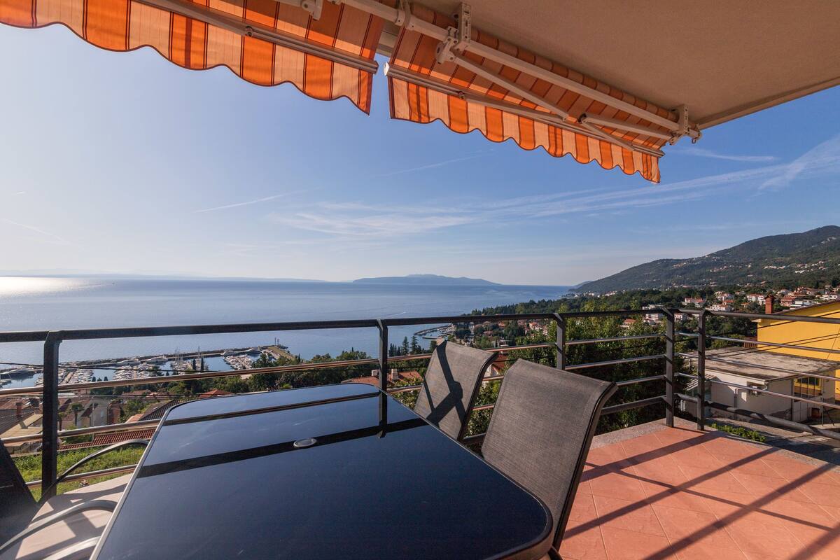 A terrace with a table and chairs, overlooking a coastal view with a body of water and distant mountains.