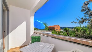 Un balcón con una mesa y sillas blancas, una sombrilla azul y vistas a una casa roja y follaje.