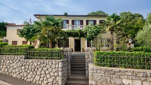 Une maison à deux étages avec une entrée en pierre, un balcon et un jardin agrémenté de palmiers.