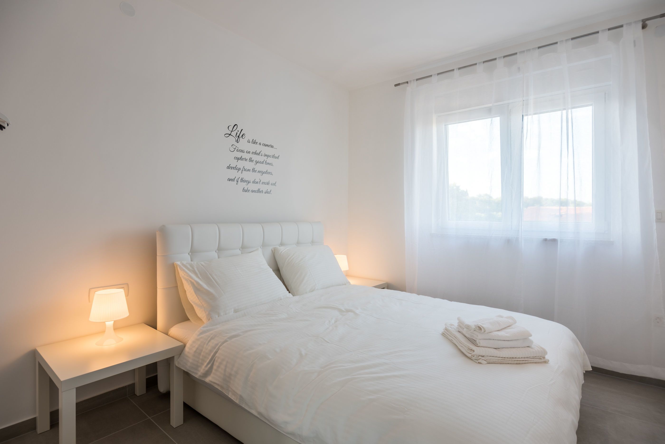 A bedroom with a bed, a bedside table, a lamp, and a window with sheer curtains.