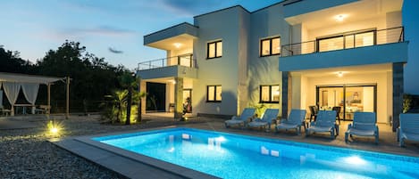 A modern two-story house with a swimming pool, outdoor seating, and a well-lit entrance.