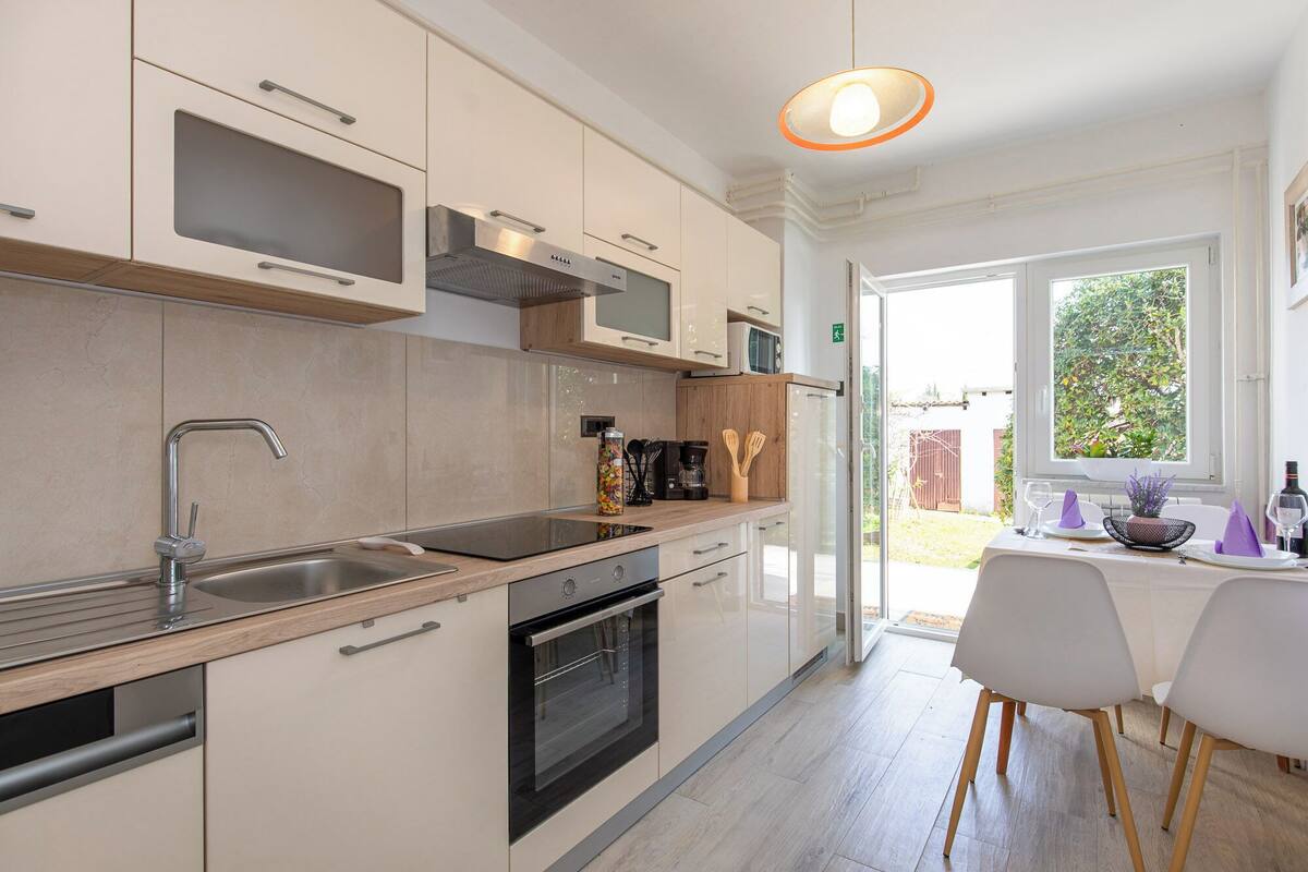 A modern kitchen with a built-in oven, microwave, and stainless steel sink. There is a dining area with a table and chairs, and a window providing a view of the outdoors.